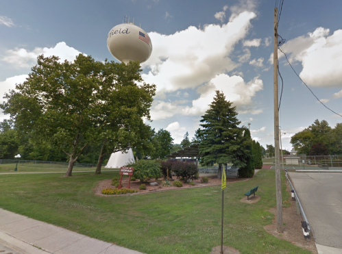 Water tower above a park with trees and shrubs, under a partly cloudy sky. A bench is visible in the foreground.