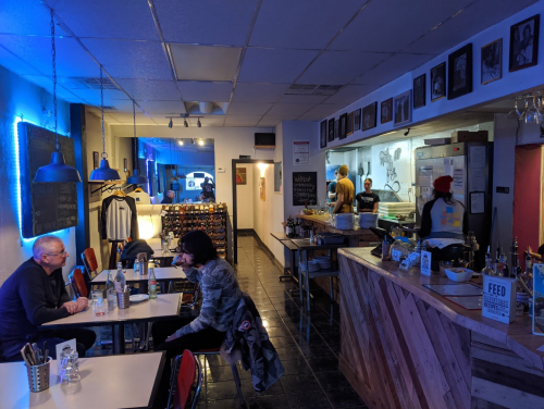 A cozy restaurant interior with diners at tables, a counter with food preparation, and blue lighting accents.