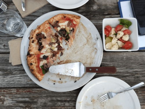 A partially eaten pizza with mushrooms and toppings on a rustic wooden table, alongside a small salad and utensils.