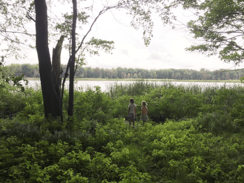 Two people stand by a lush green area near a calm lake, surrounded by trees and tall grasses under a cloudy sky.