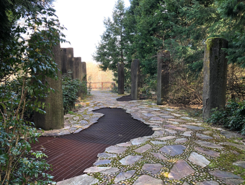 A stone pathway surrounded by greenery, featuring tall stone pillars and a unique textured area on the ground.