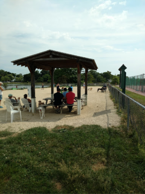 A group of people sitting under a gazebo at a park, with a sandy area and a tennis court in the background.