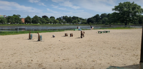 A child runs on a sandy area near a river, with playground equipment and trees in the background.