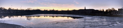 A serene lake at sunset, reflecting colorful skies and surrounding trees, with a hint of ice on the water's edge.