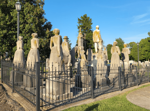 A group of weathered stone statues in a fenced area, surrounded by trees and a clear blue sky.