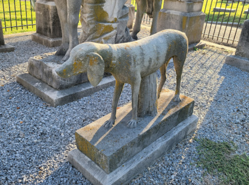 A weathered stone statue of a dog standing on a pedestal, surrounded by other sculptures in a gravel area.