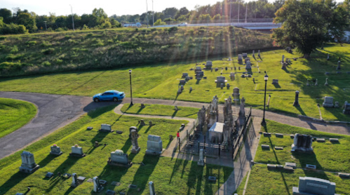 Aerial view of a cemetery with gravestones and a blue car parked on a path, surrounded by green grass and trees.