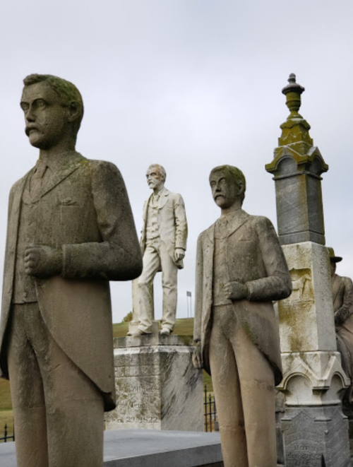 Three stone statues of men, with one larger figure in the center, set against a cloudy sky.