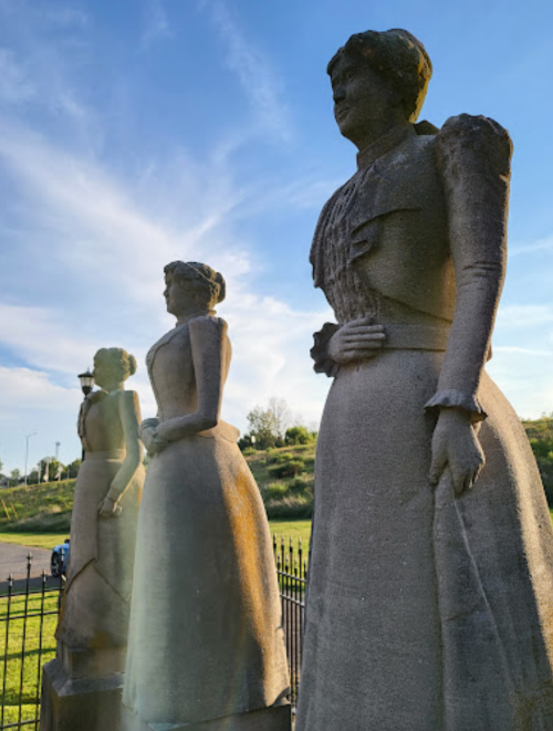 Three stone statues of women in historical dresses, set against a blue sky with wispy clouds.