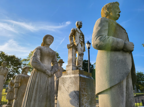 A group of stone statues, including a man on a pedestal and a woman in a dress, set against a blue sky.