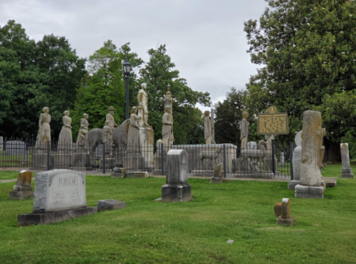 A cemetery with various stone statues and gravestones, surrounded by greenery and a historical marker.