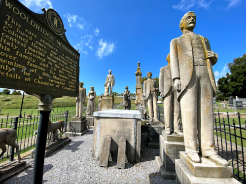 A historic cemetery with statues and a plaque, featuring the Wooldridge Monument and surrounding greenery under a blue sky.