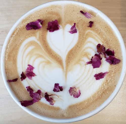 A latte with intricate heart-shaped foam art, topped with scattered rose petals.
