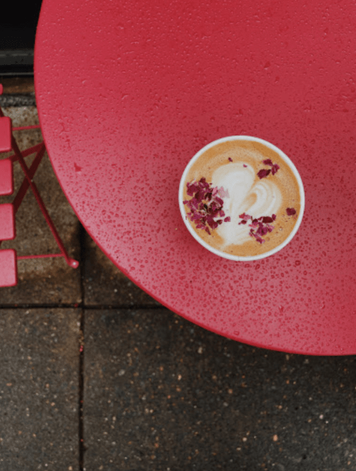A top-down view of a latte with rose petals on a bright red table, surrounded by raindrops.