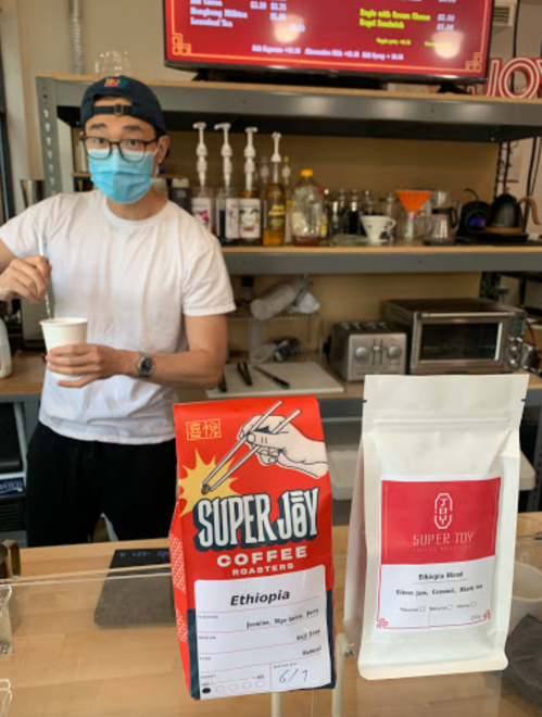 A barista in a mask prepares coffee at a counter, with bags of Super Joy coffee on display.