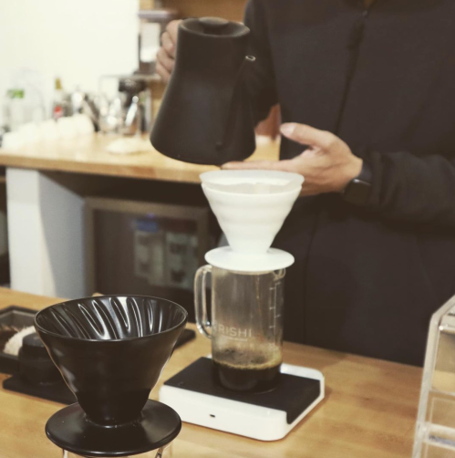 A person pours water from a kettle into a coffee dripper set over a blender, preparing coffee in a cozy kitchen.
