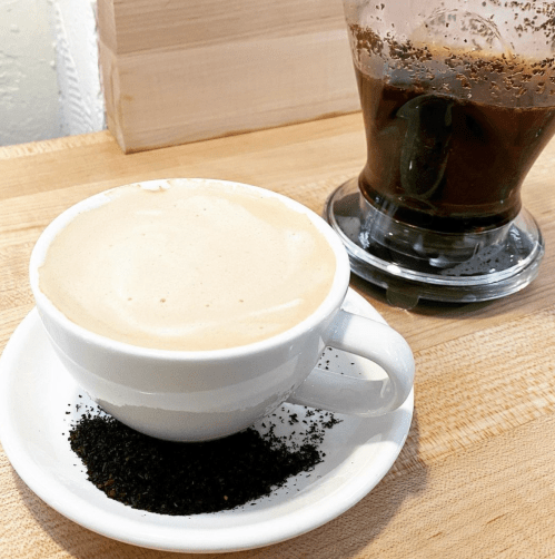 A cup of coffee with frothy milk sits on a saucer with coffee grounds, next to a glass of dark liquid.