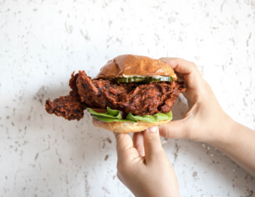 A person holding a crispy fried chicken sandwich with lettuce and pickles on a textured background.