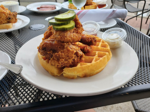 A plate featuring fried chicken on a waffle, topped with pickles and served with dipping sauces.