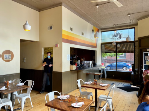 A cozy restaurant interior with wooden tables, white chairs, and a staff member wearing a mask walking through.