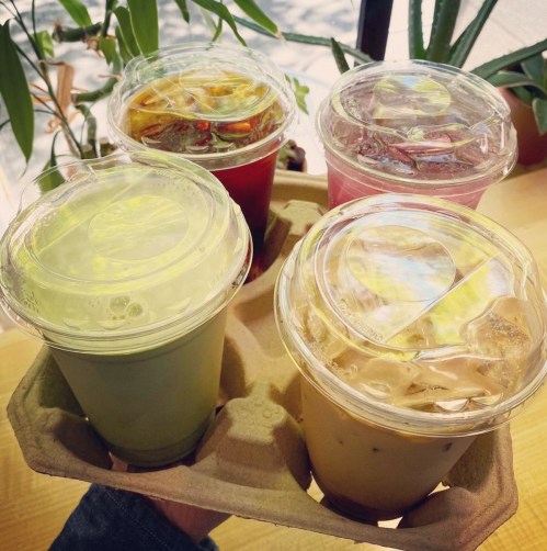 Four colorful drinks in clear cups, held in a cardboard carrier, with greenery in the background.