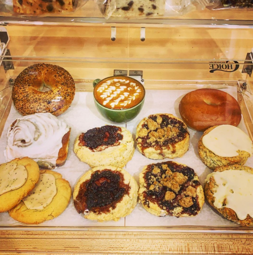 A display of assorted baked goods, including bagels, cookies, and a latte art coffee in a green cup.