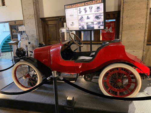 A vintage red 1914 Saginaw cyclecar displayed in a museum, featuring whitewall tires and a classic design.