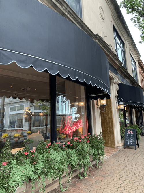 A storefront with black awnings, neon sign reading "Lit & Inspired," and flower-filled planters outside.