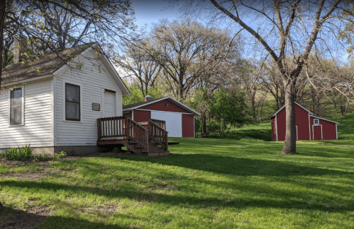 A white house with a wooden deck surrounded by green grass, with two red barns in the background and trees nearby.