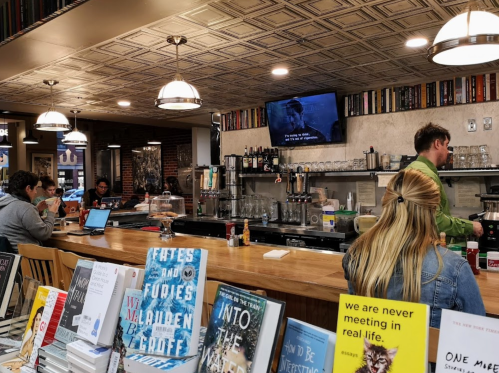 A cozy café with a bar, bookshelves, and patrons working on laptops, while a movie plays on a screen in the background.