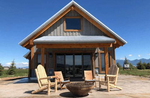 A cozy wooden cabin with a metal roof, featuring outdoor seating and a fire pit, set against a clear blue sky.