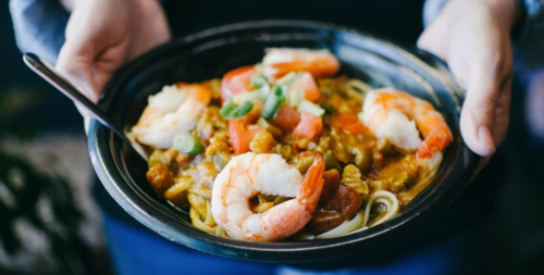 A person holding a bowl of shrimp pasta topped with green onions and colorful vegetables.
