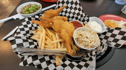 A basket of fried shrimp with fries, coleslaw, and dipping sauces on a checkered paper.