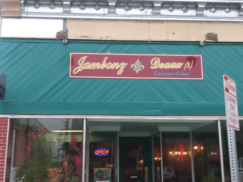 Sign for "Jambony Deaux" featuring a fleur-de-lis, with a colorful storefront and "Open" sign visible.