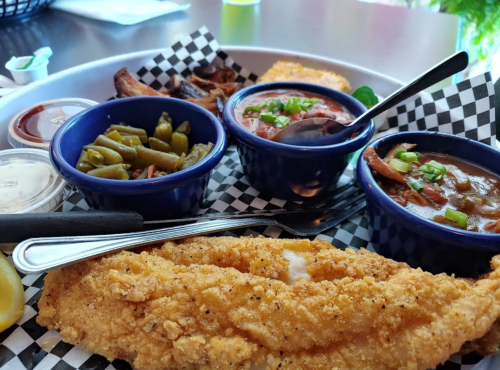 A plate featuring fried fish, green beans, gumbo, and a side of sauce, served with lemon wedges on a checkered tablecloth.