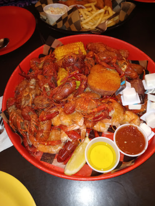 A red platter filled with crawfish, shrimp, corn, sausage, and dipping sauces, served with lemon and hushpuppies.