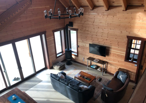 Cozy wooden living room with a sofa, armchair, coffee table, and a TV, featuring large windows and a rustic chandelier.