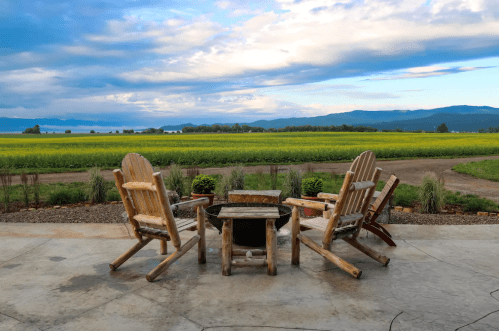 A scenic view of a patio with wooden chairs and a table, overlooking a lush green field and distant mountains.