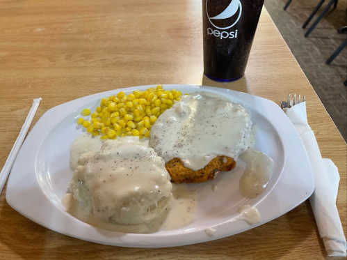 A plate of fried chicken with gravy, mashed potatoes, corn, and a Pepsi drink on a wooden table.