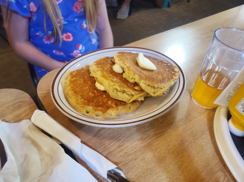 A plate of three golden pancakes topped with butter, sitting on a wooden table next to a glass of drink.