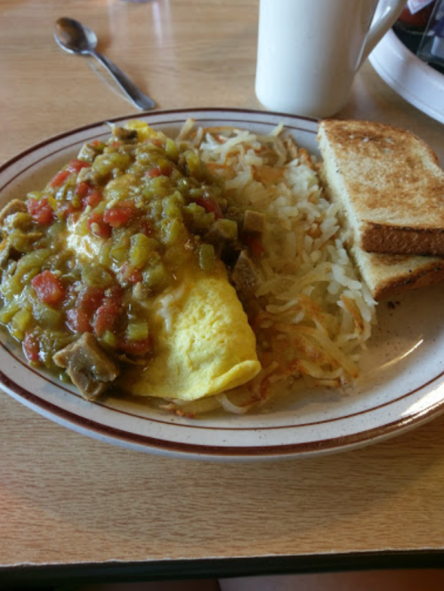 A plate with an omelet topped with green chili, hash browns, and toast, alongside a cup of coffee.