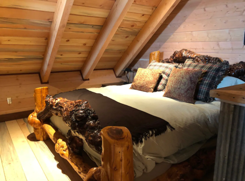 Cozy attic bedroom with a wooden bed, decorative pillows, and sloped wooden ceiling. Warm and inviting atmosphere.