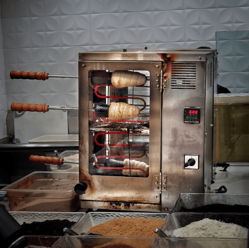 A vertical rotisserie with cooking meat, surrounded by various ingredients in containers on a countertop.