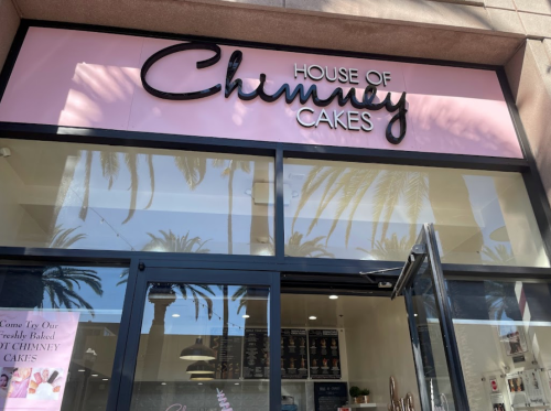 Sign for "House of Chimney Cakes" with palm trees reflected in the glass entrance.