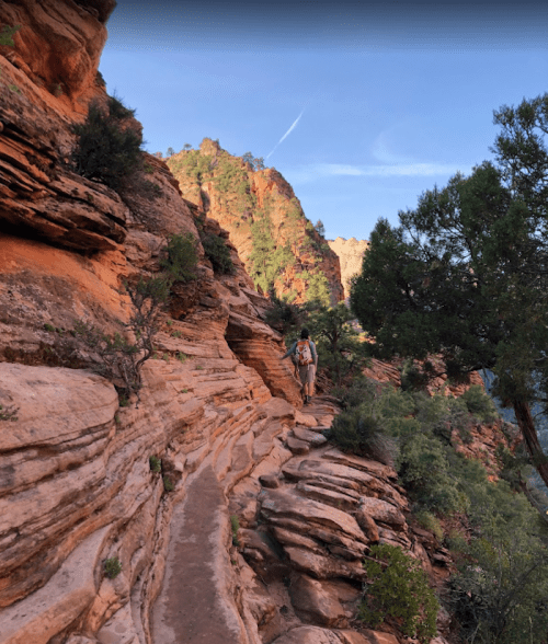 A hiker walks along a narrow, rocky trail surrounded by red rock formations and greenery under a blue sky.