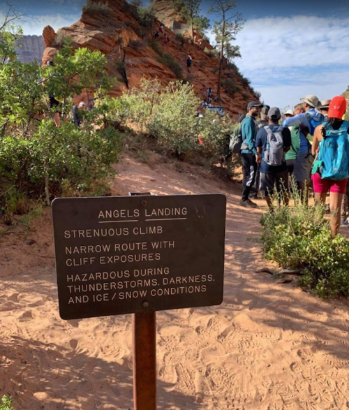 Sign for Angels Landing trail, warning of strenuous climb and hazards like thunderstorms and icy conditions, with hikers in background.