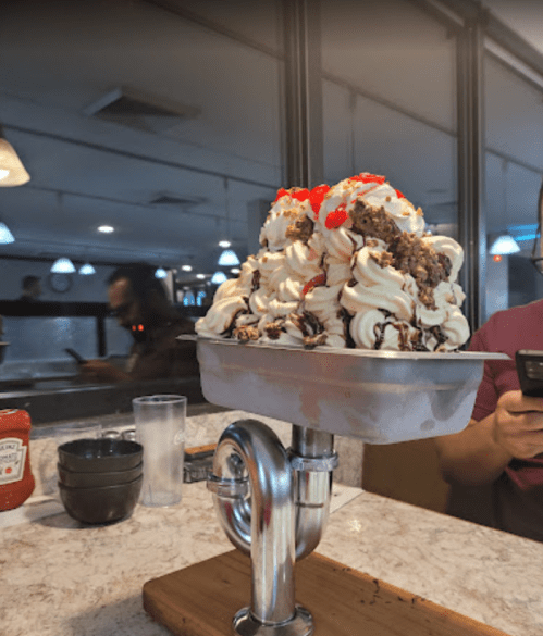 A large dessert with whipped cream, cherries, and chocolate sauce served in a metal dish on a wooden base.