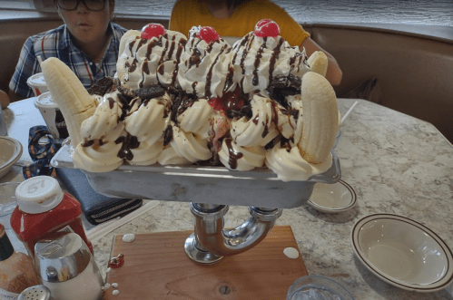 A large ice cream sundae with bananas, whipped cream, chocolate drizzle, and cherries, served in a metal dish.