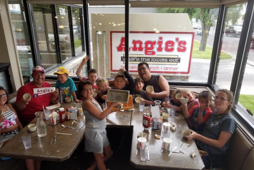A group of children and adults enjoying a meal at Angie's restaurant, smiling and holding empty bowls at a table.