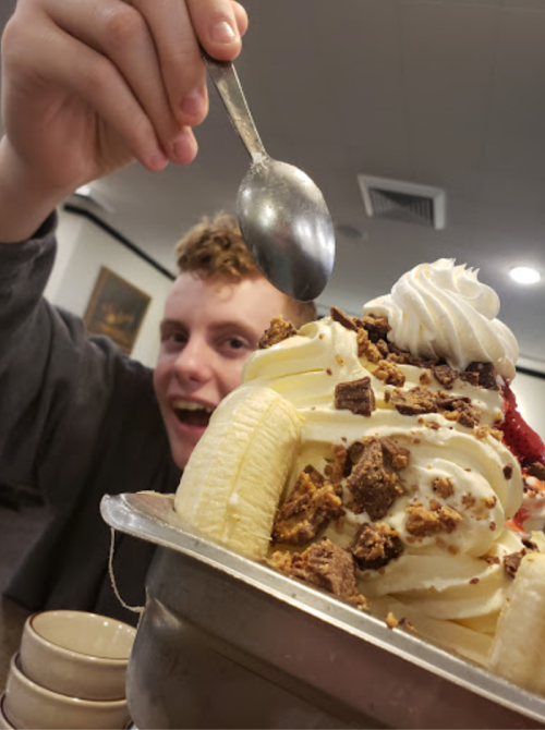 A person holds a spoon above a large dessert topped with whipped cream, banana slices, and crushed toppings.